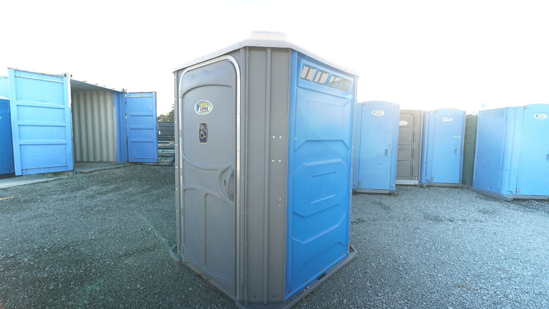 A gray and blue portable toilet stands among other similar units on gravel.