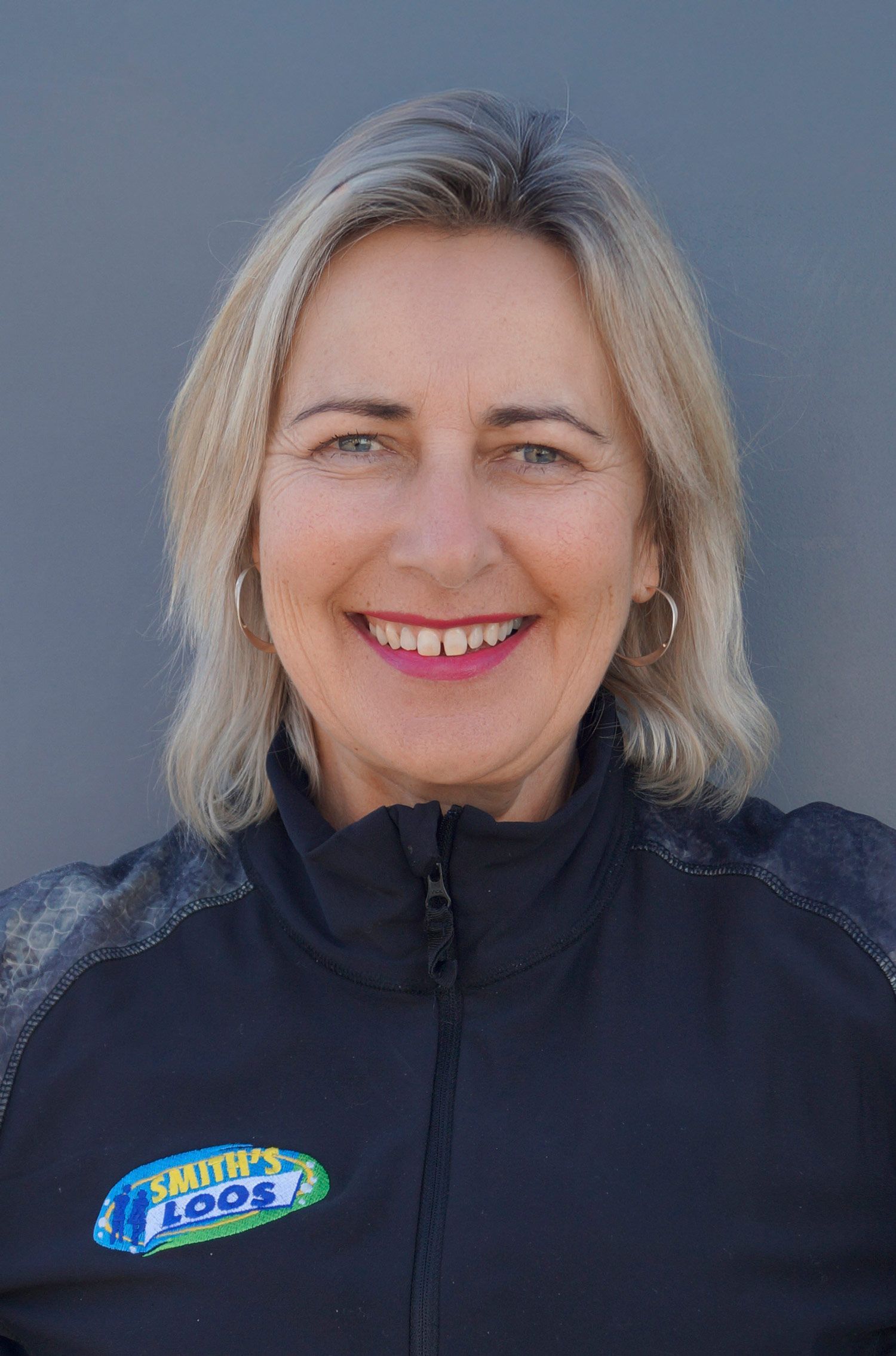 Blonde woman smiling, wearing a navy jacket with a logo. Against a plain gray background.