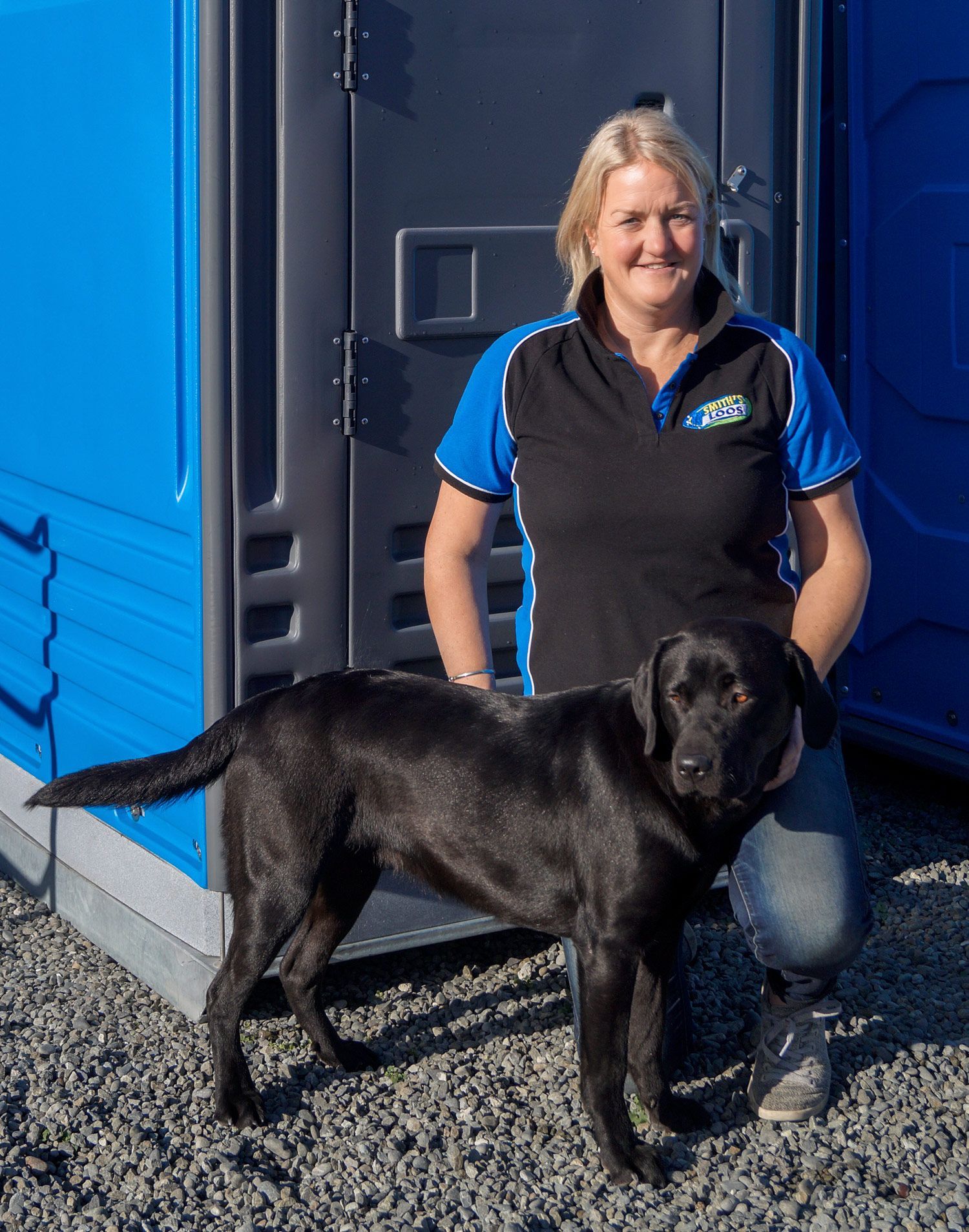 Woman and black Labrador standing beside blue portable toilets.
