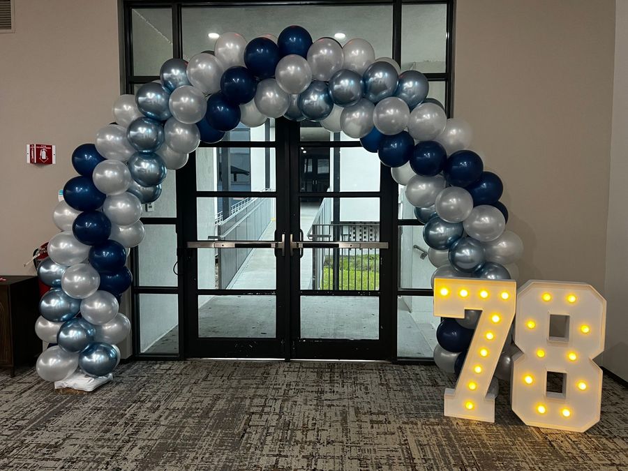 Silver and blue balloon arch beside lit “78” numbers at a doorway.