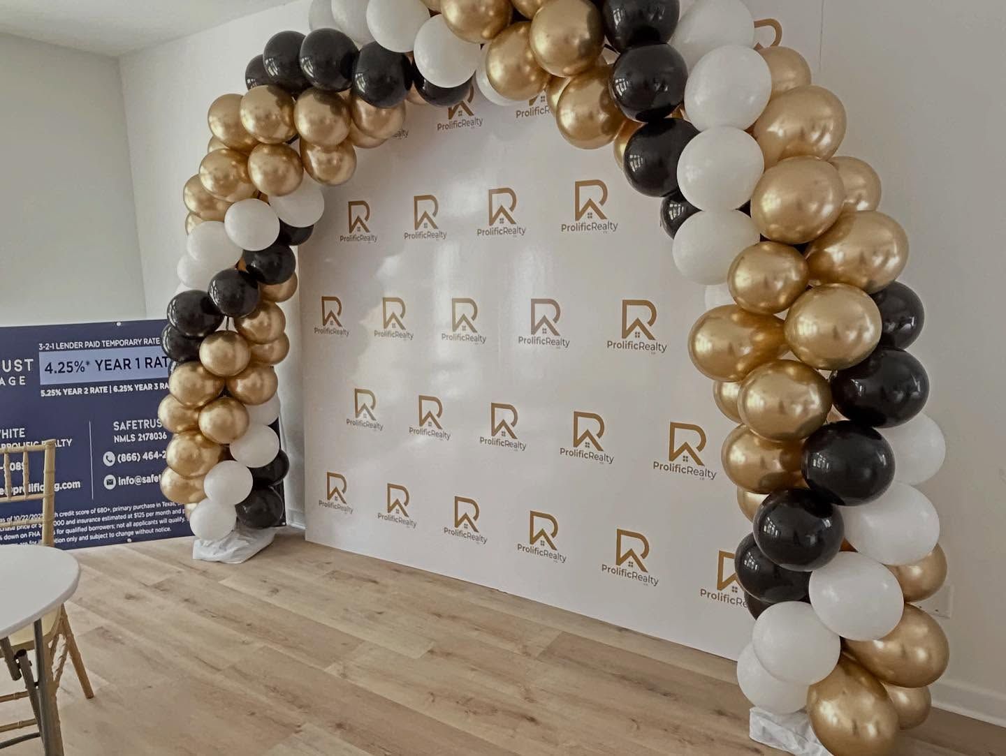 Gold, black, and white balloon arch at an event backdrop with logo pattern on a wooden floor