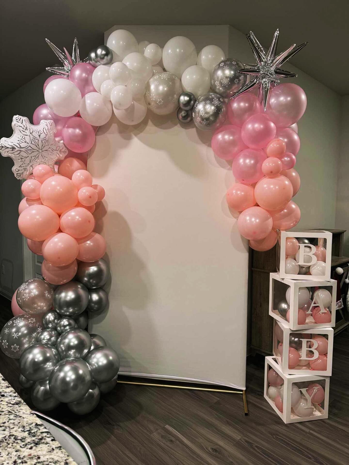 Pink, white, and silver balloon arch framing a doorway, with stacked gift boxes on the right.