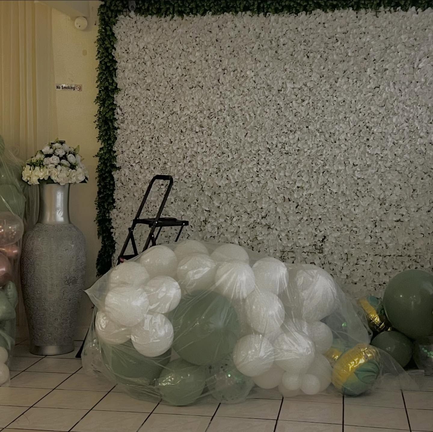 Floral backdrop with white and green balloons on the floor beside a tall vase of flowers and a folded chair