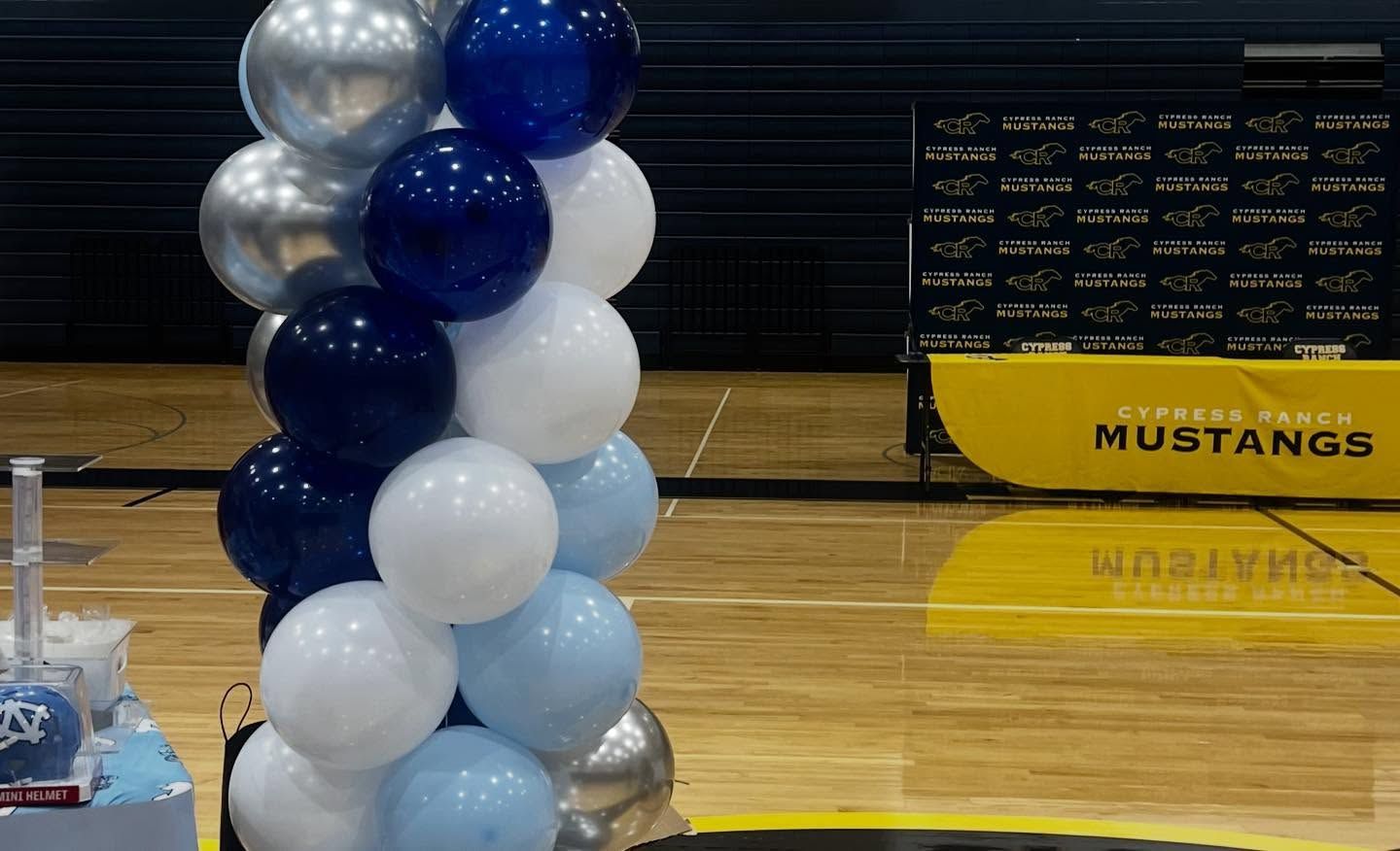 Blue, white, and silver balloon column on a gym floor with Mustangs bleachers in the background