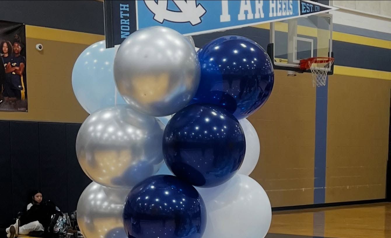 Cluster of metallic blue, silver, and white balloons in a gymnasium near a basketball hoop.