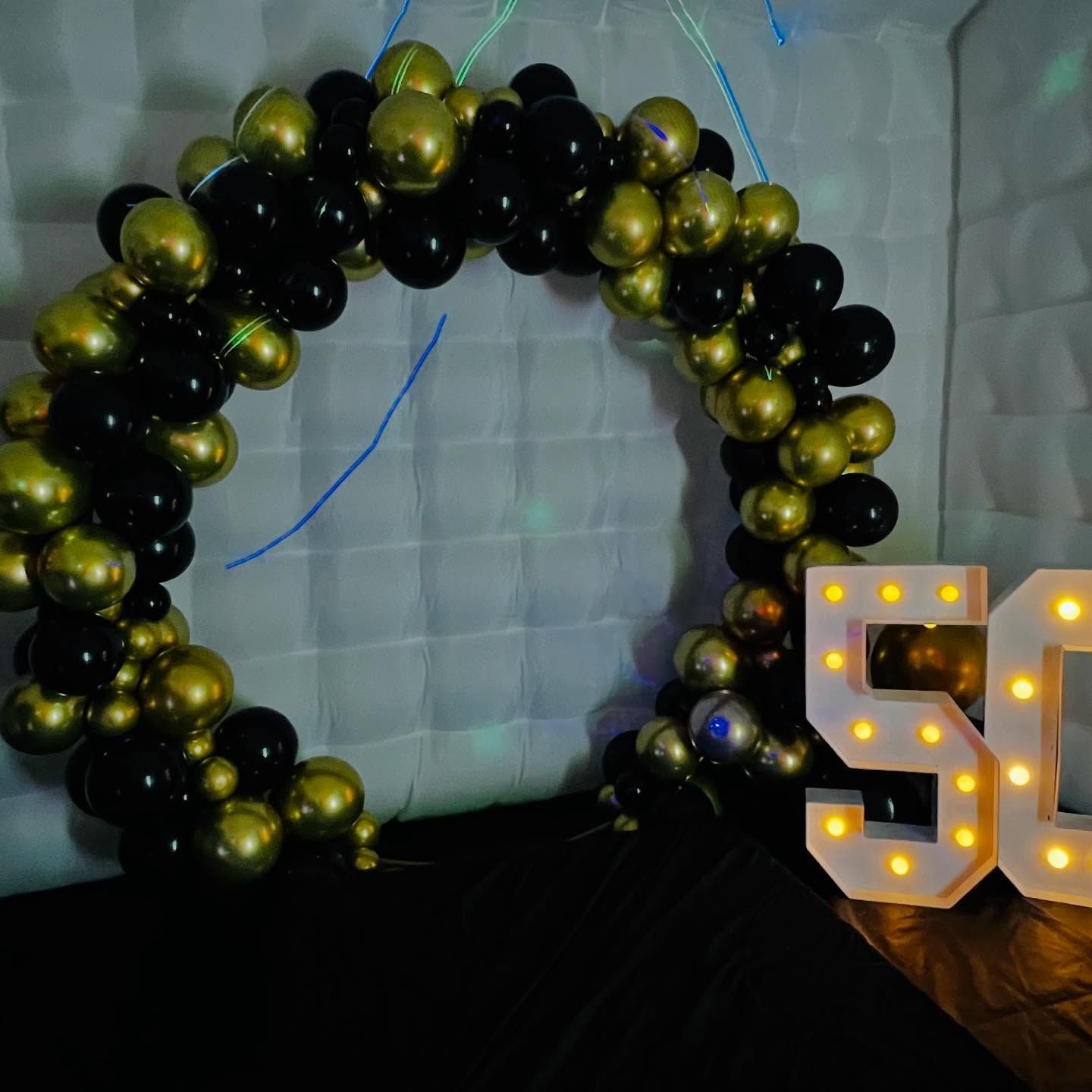 Black and gold balloon arch beside illuminated “50” numbers on a white backdrop