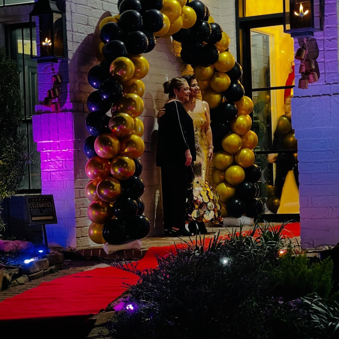 Couple posing under black-and-gold balloon arch on a red carpet at a nighttime event