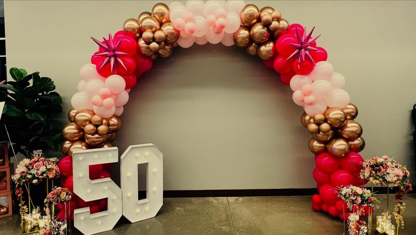50th birthday balloon arch with red, gold, and blush balloons and large white 