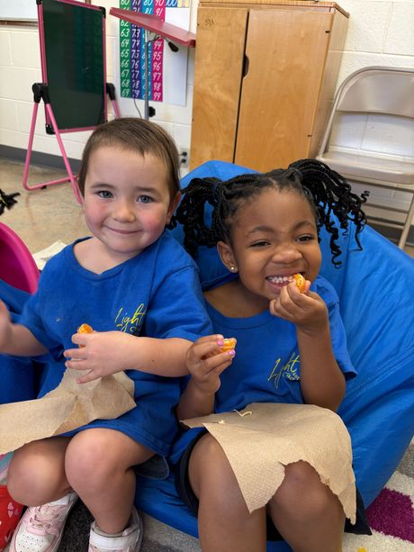 Two children in blue shirts smiling and eating snacks on a blue beanbag chair. Classroom setting.