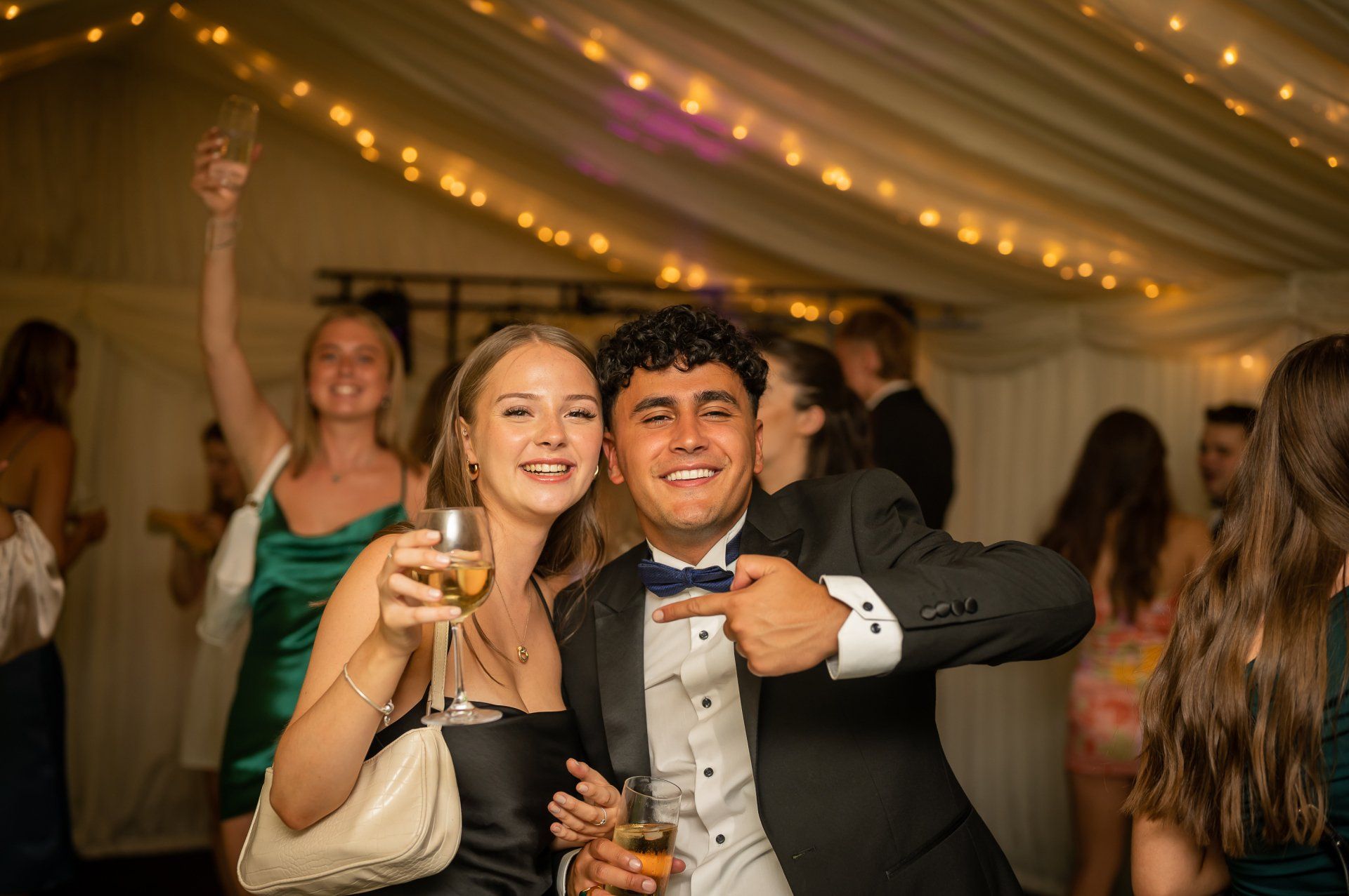 Image of people at a formal gathering which was a 21st birthday party with a girl & young man in the foreground each holding drinks.