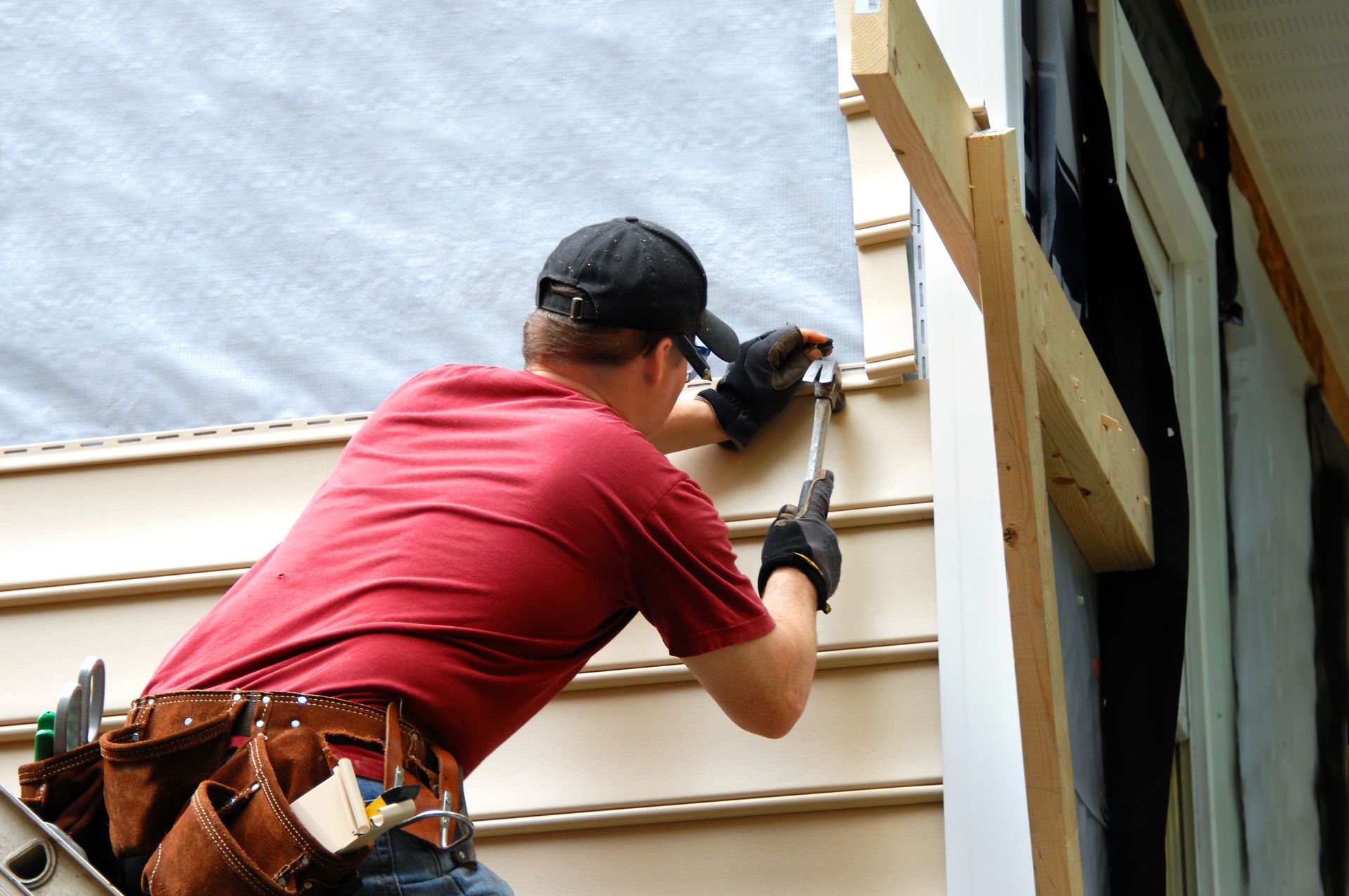 Person in red shirt and cap installing siding on a house; using hammer.