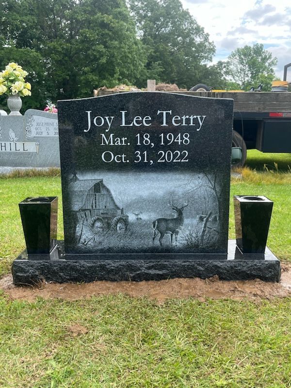 Black granite headstone with name, dates, and etched scene of barn and deer. Two black vases flank it.