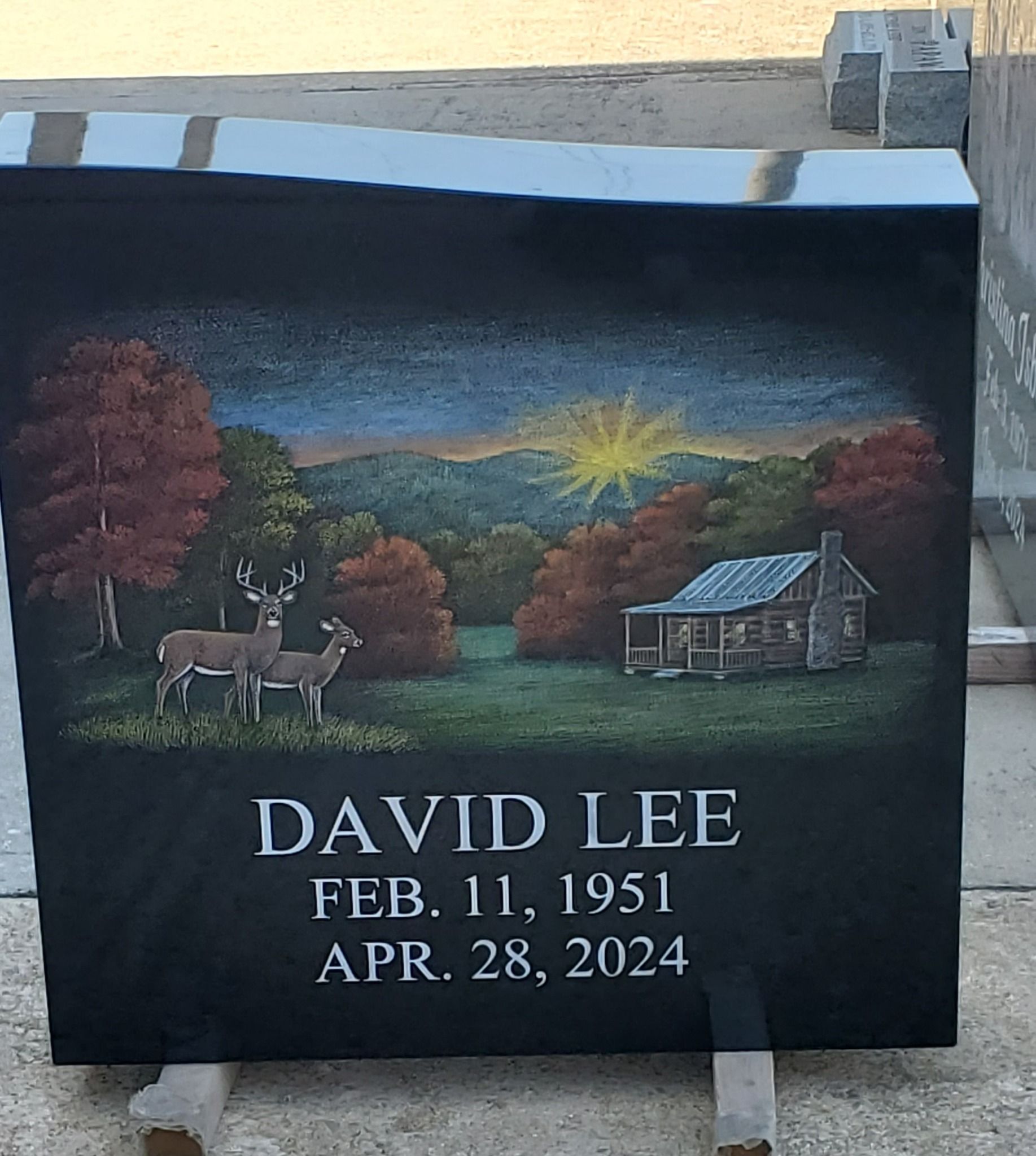 Black headstone with landscape painting: cabin, deer, sunrise, name, and dates.