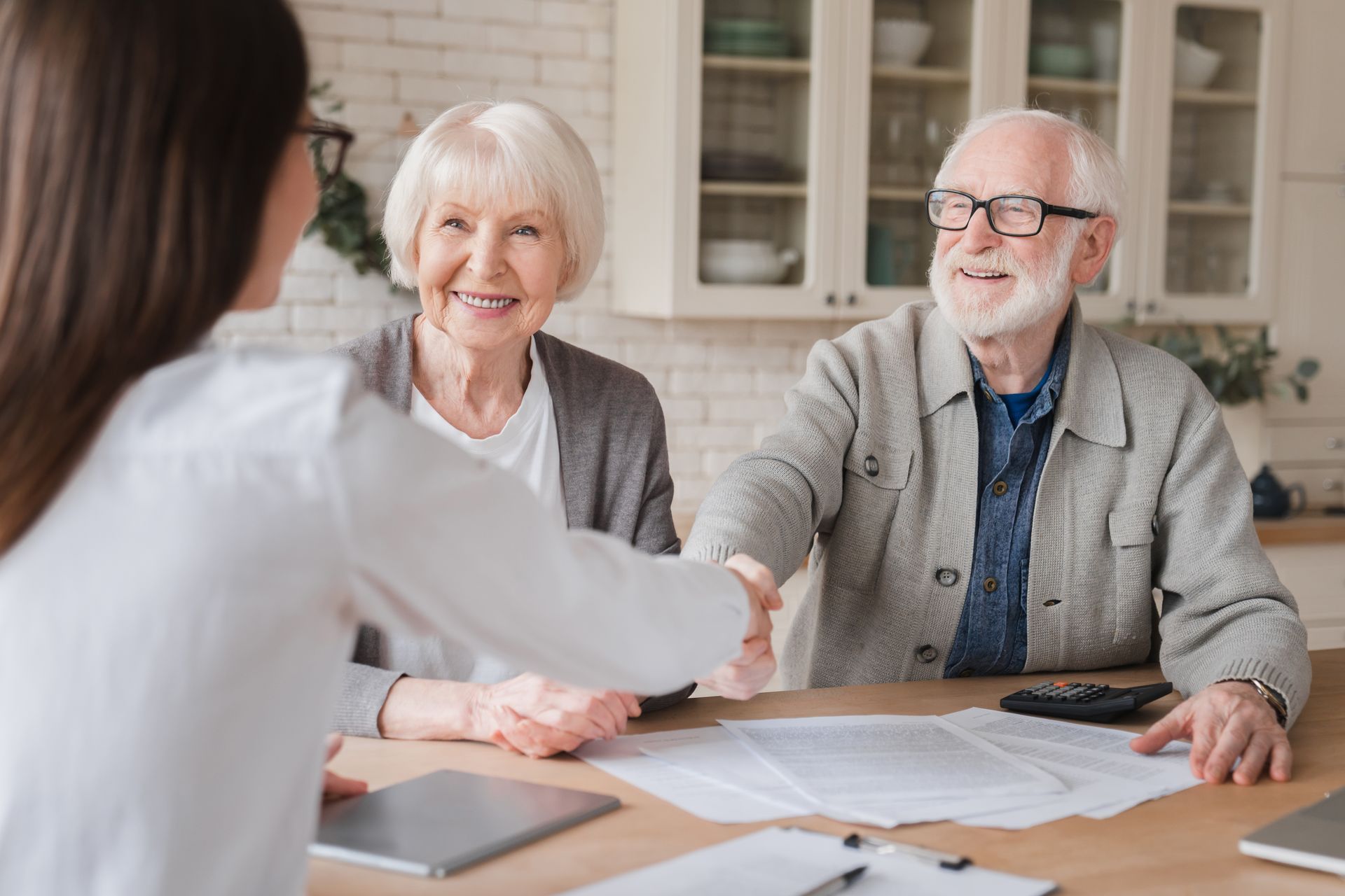 Woman shaking hands with a smiling older man; another woman sits nearby. Papers and a calculator are on the table in a kitchen.