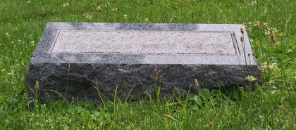 A rectangular, gray granite headstone lies flat on a grassy lawn.