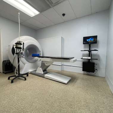 Medical imaging room with PET scanner and bed. Equipment on the left, a computer screen on the right, light tan floor.