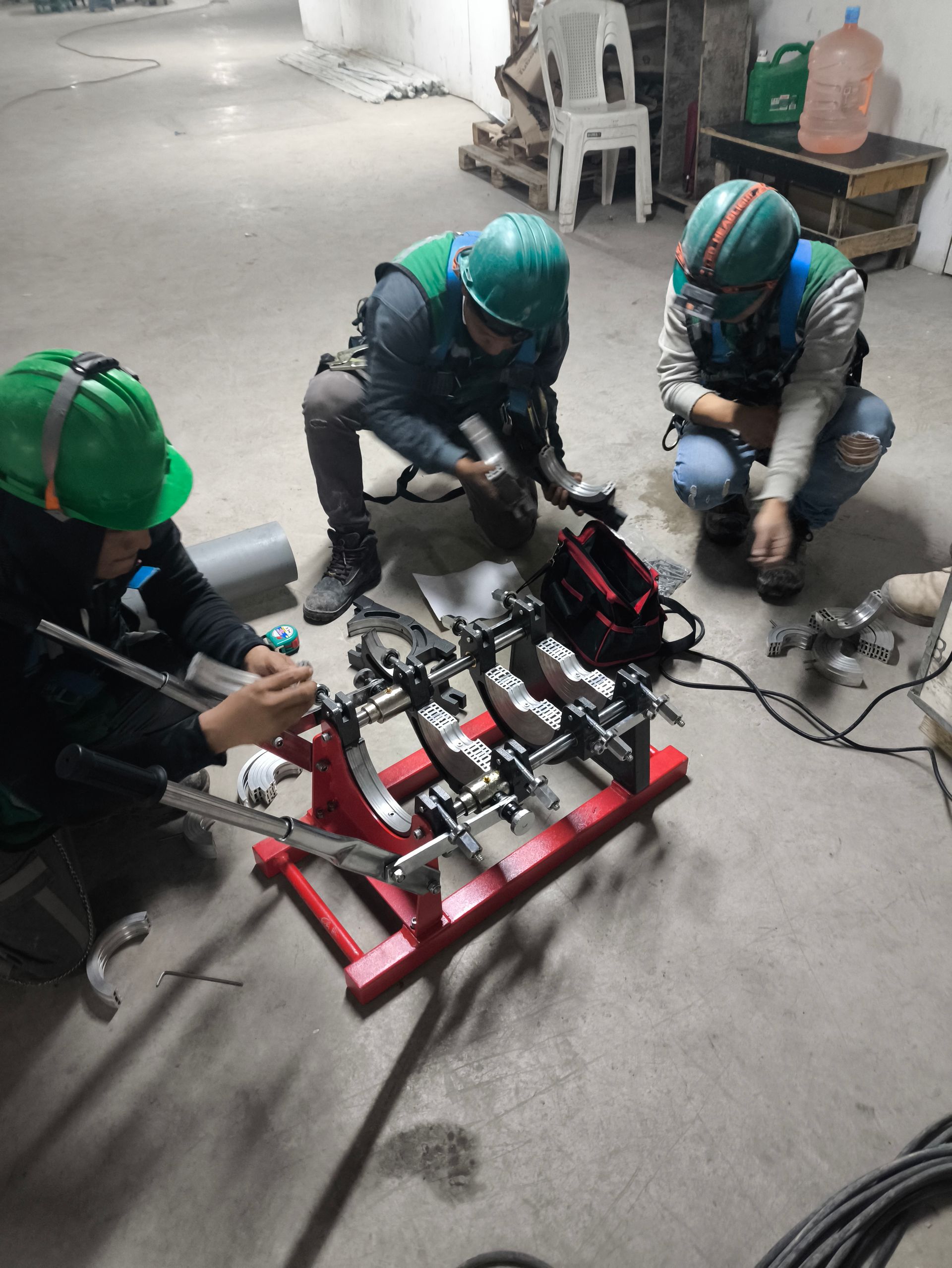 Tres trabajadores con cascos utilizan herramientas para ensamblar tuberías en un soporte rojo en un taller.