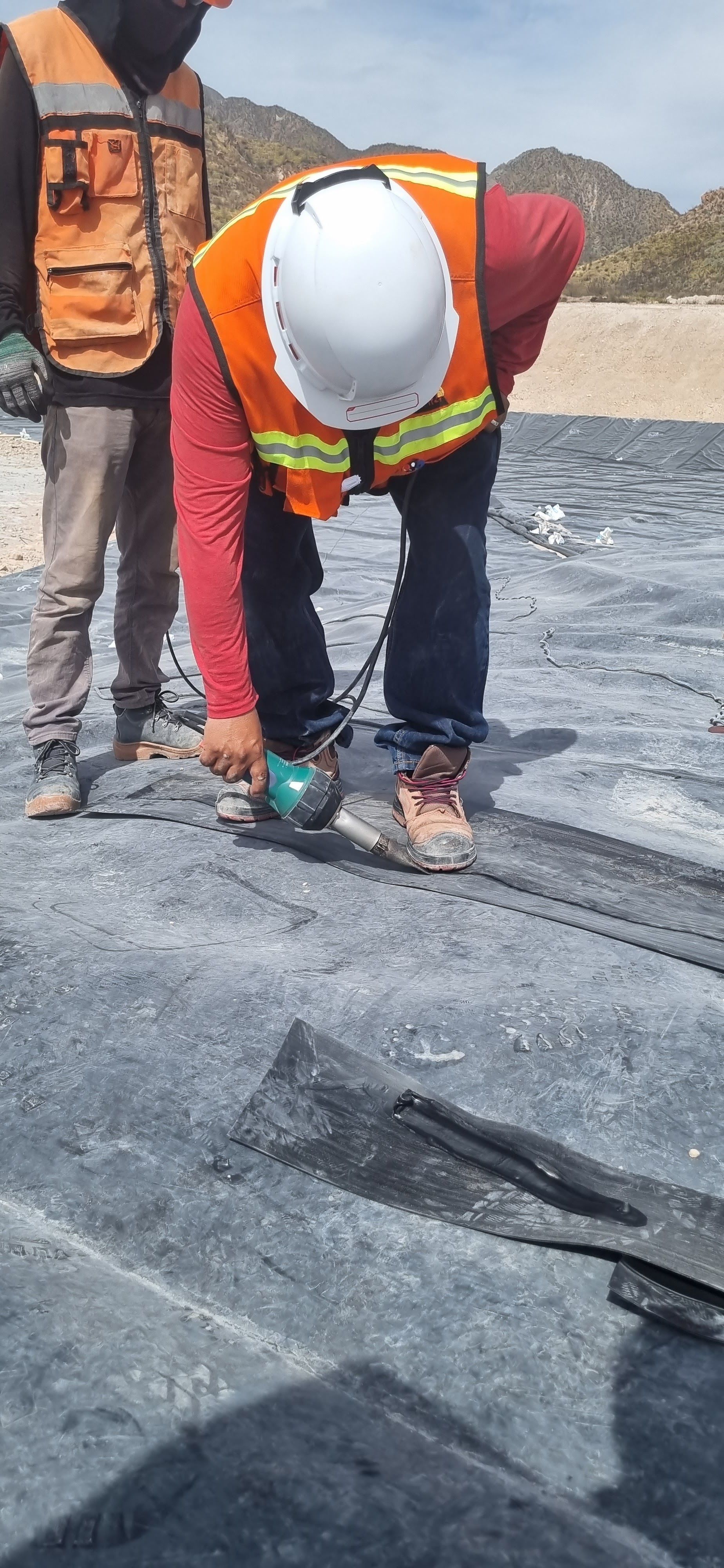 Dos trabajadores cortando un material oscuro con una herramienta eléctrica, uno de ellos con chaleco de seguridad y casco.