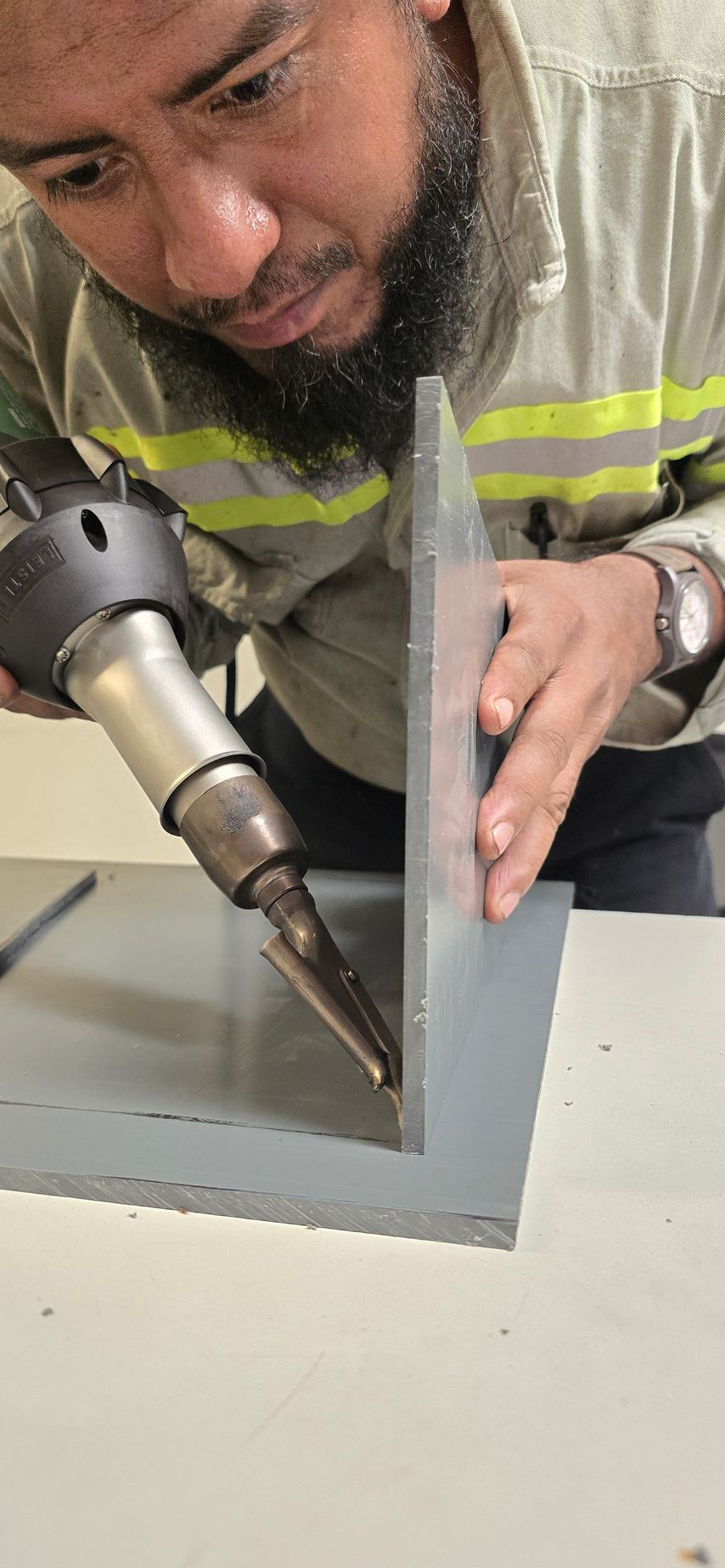 Un hombre usa una pistola de calor para soldar dos paneles grises en una esquina. Lleva chaleco de seguridad.