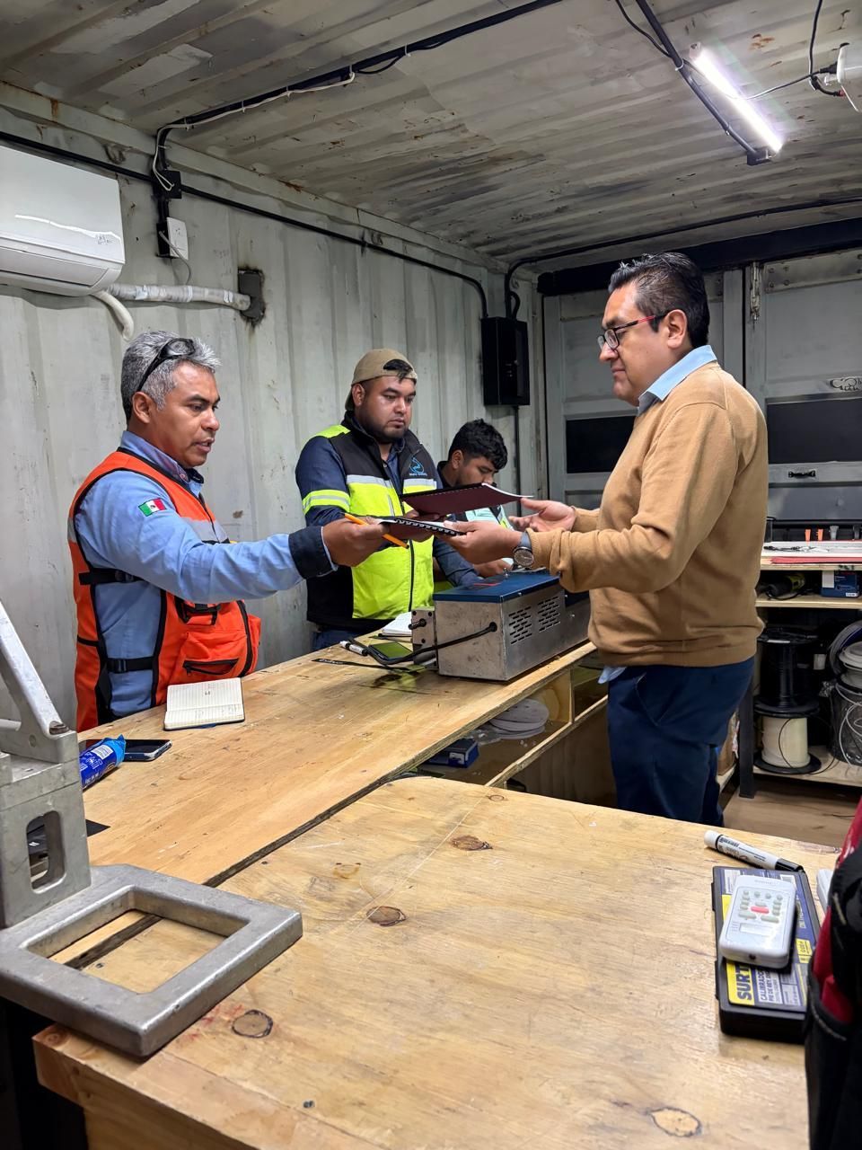 Tres hombres intercambian documentos en una pequeña oficina industrial. Un hombre con chaleco naranja entrega papeles a otro con suéter.