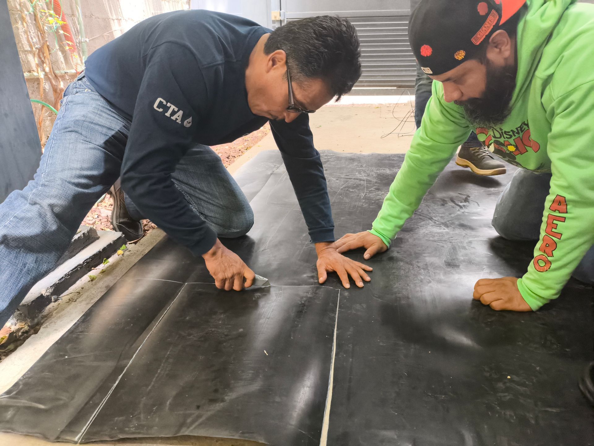 Dos hombres arrodillados, cortando y colocando una lámina de tela negra en el suelo de un interior.
