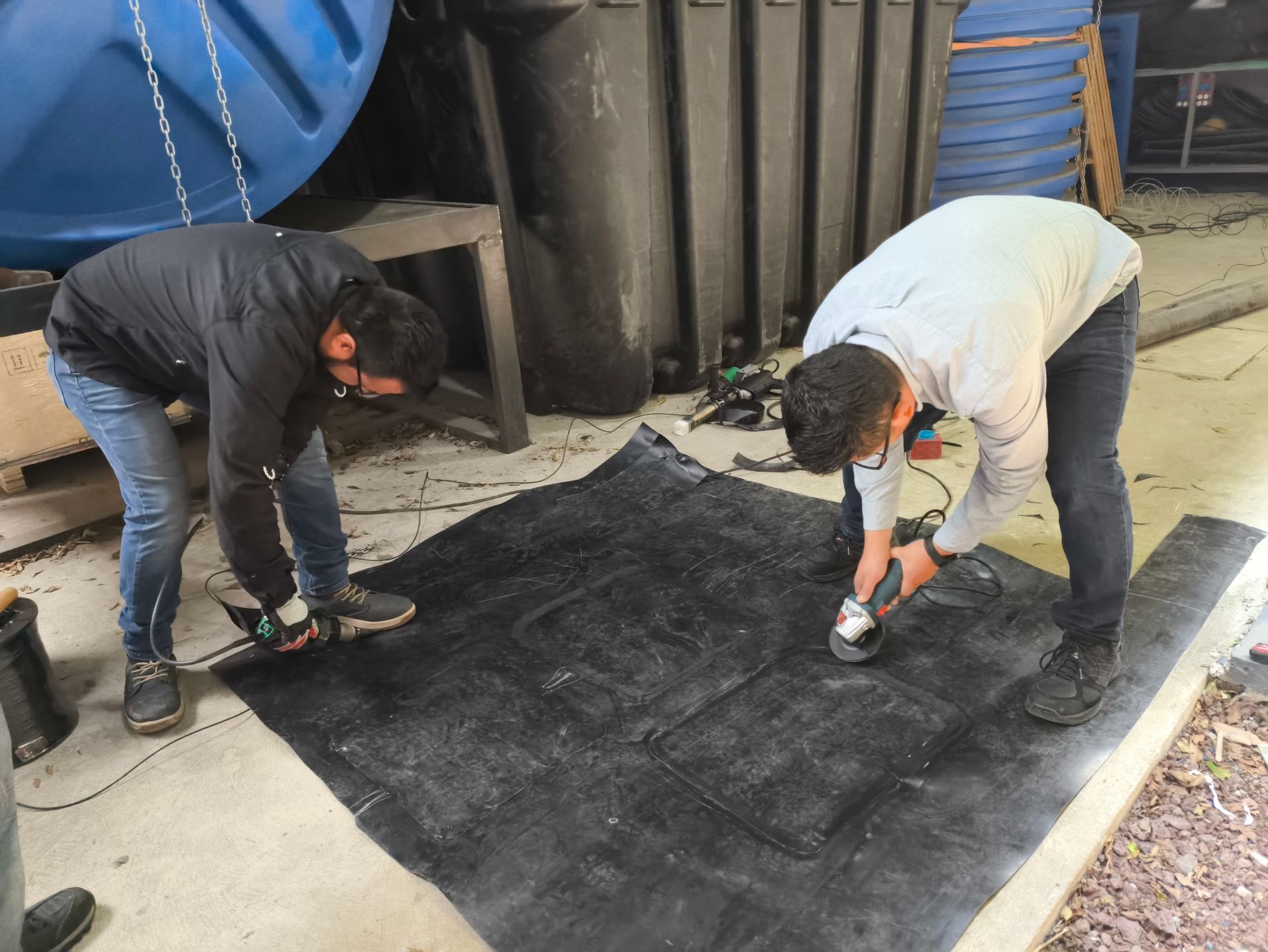 Dos personas cortan una alfombra negra sobre una superficie de concreto con herramientas eléctricas cerca de tanques y contenedores azules.