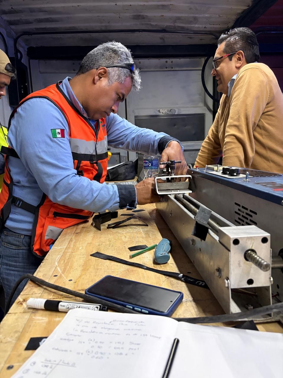 Un hombre con chaleco naranja repara maquinaria mientras otro hombre lo observa. Dentro de un taller, con herramientas y un cuaderno a la vista.