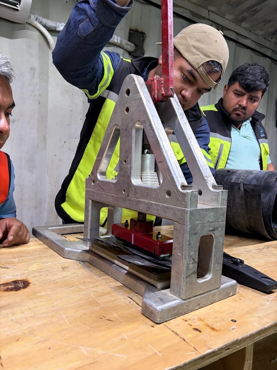 Un trabajador usa una prensa manual, con otros dos hombres observándolo. Dentro de una habitación, con una mesa de madera.
