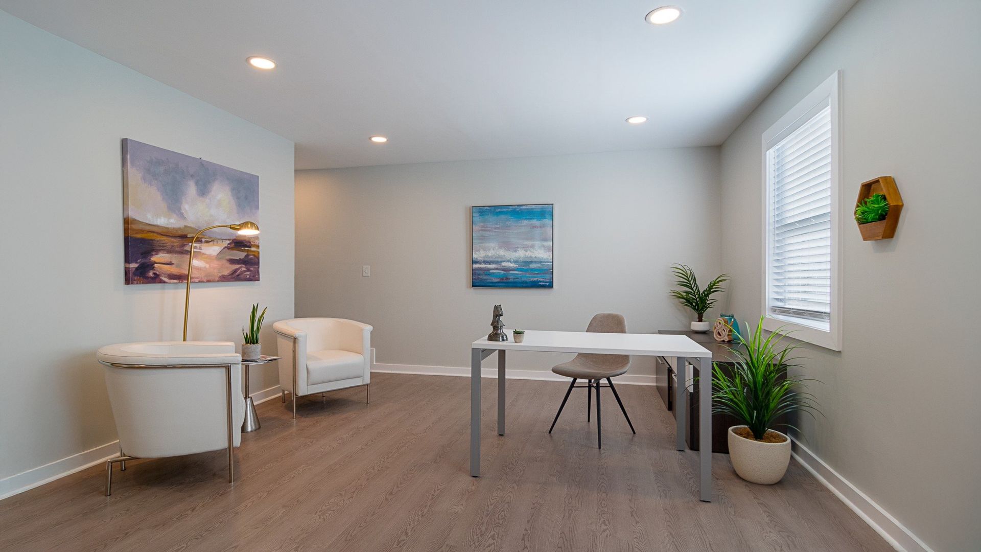 Bright minimalist home office with desk, chairs, wall art, and potted plants by a window.