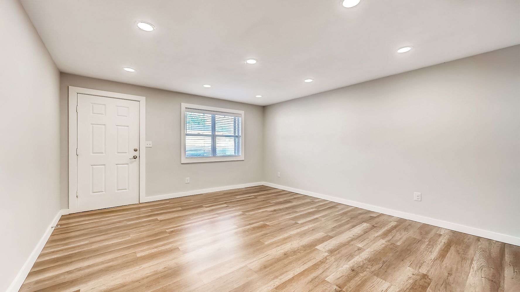 Empty beige room with wood floors, white walls, recessed lights, and a small window.