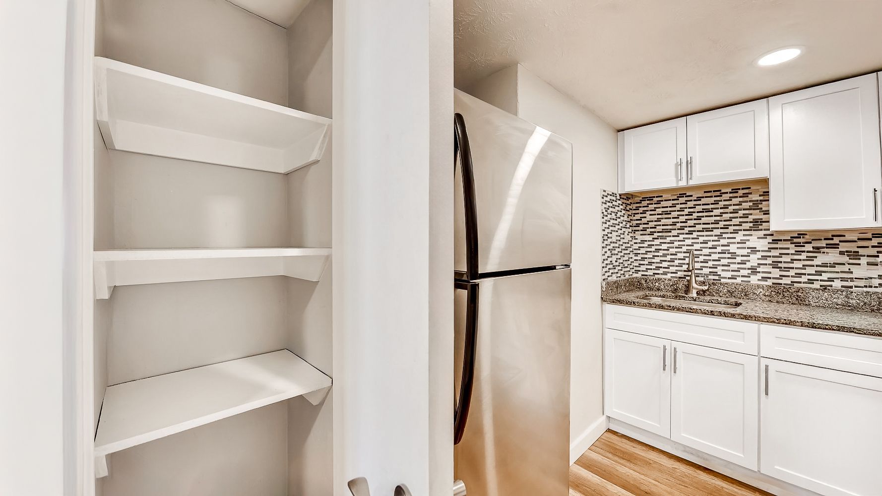 Pantry shelves beside a white kitchen with cabinets, mosaic backsplash, and hardwood floor
