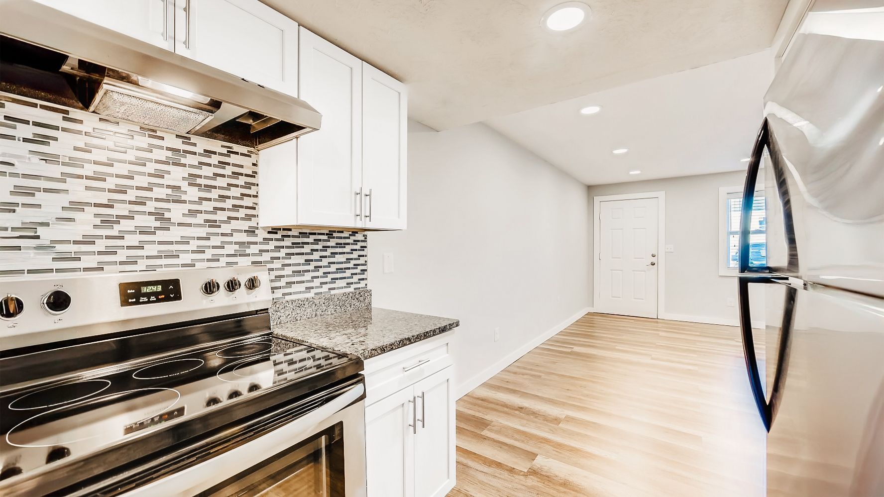 Bright kitchen with white cabinets, stainless steel stove, tiled backsplash, and light wood flooring leading to a hallway