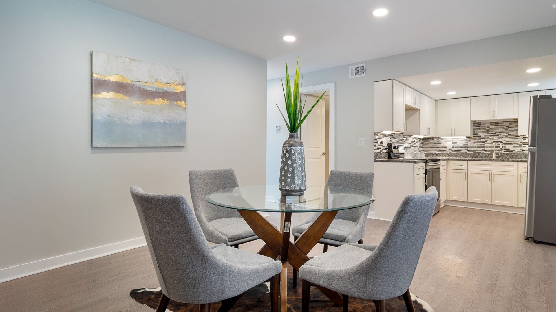 Modern dining area with glass table, gray chairs, wall art, and open view into a bright kitchen