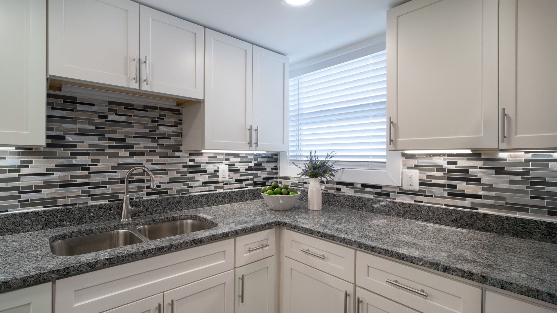 Bright modern kitchen with white cabinets, gray mosaic backsplash, granite counters, and a window over the sink