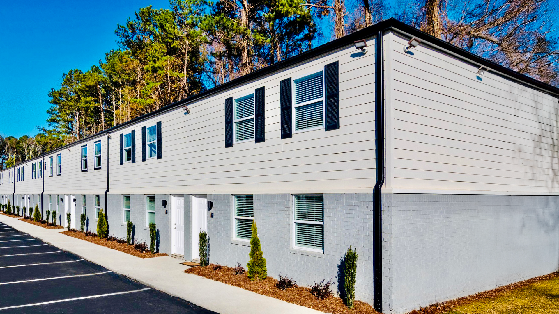 Two-story white apartment building with dark shutters beside a parking lot and trees