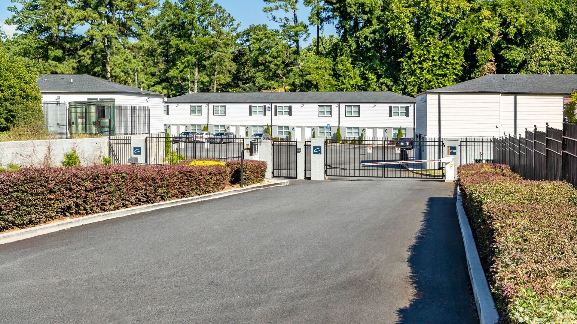 Modern white building with a gated entrance, paved driveway, and hedges on both sides