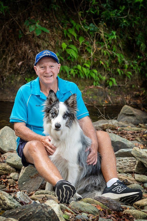 Man in blue shirt and baseball cap sits with a blue merle Border Collie on rocks near a stream.— Canine Training School in Trinity Park, QLD