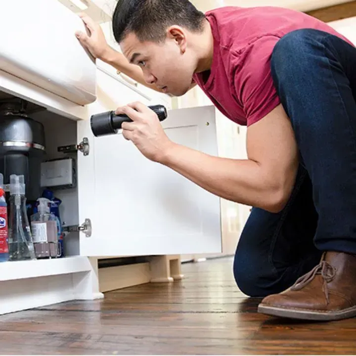 Man examines under-sink cabinet with flashlight, focused. Red shirt, jeans, and brown shoes.