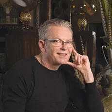 Man with gray hair, glasses, and a black shirt smiles, resting his hand near his face.