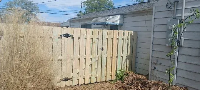 Wooden fence in front of a house, with a gate and a patch of tall grass.