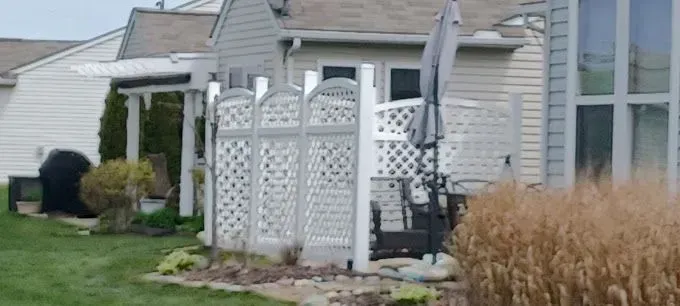 Backyard patio with white lattice fence, umbrella, and chairs. Beige siding, green lawn.