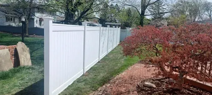 White vinyl fence bordering a yard with green grass, red bushes and trees in the background.