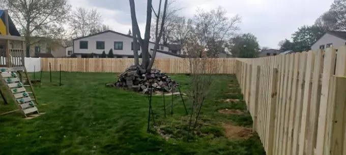 A backyard with a newly built wooden fence, trees, and green grass. A play set is in the yard.