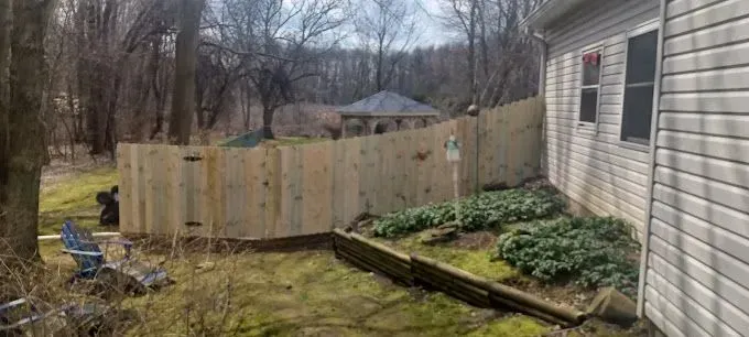 Wooden fence in a yard next to a house with greenery and trees in the background.