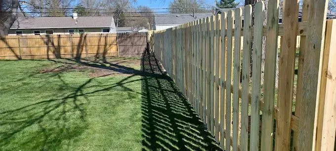 Wooden fence in a backyard casting a long shadow on the green grass. Houses and trees are in the background.