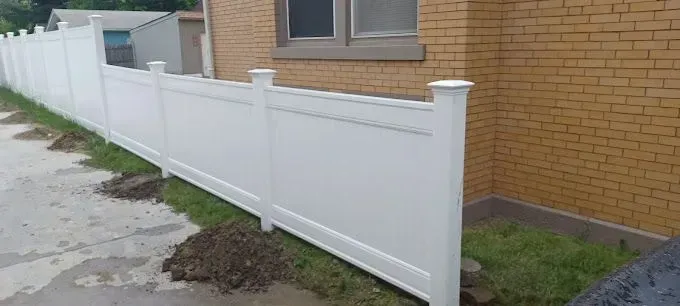 White vinyl fence alongside a yellow brick building, small shed in the background. Grass and dirt.