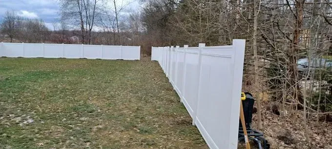 A white vinyl fence runs along a grassy area, in front of a tree line on an overcast day.