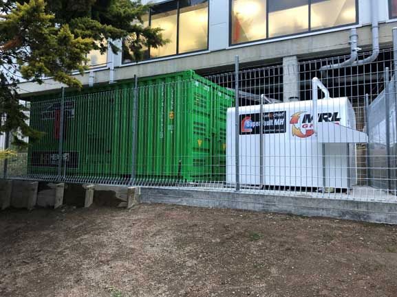 A green container and a white container are behind a fence in front of a building.