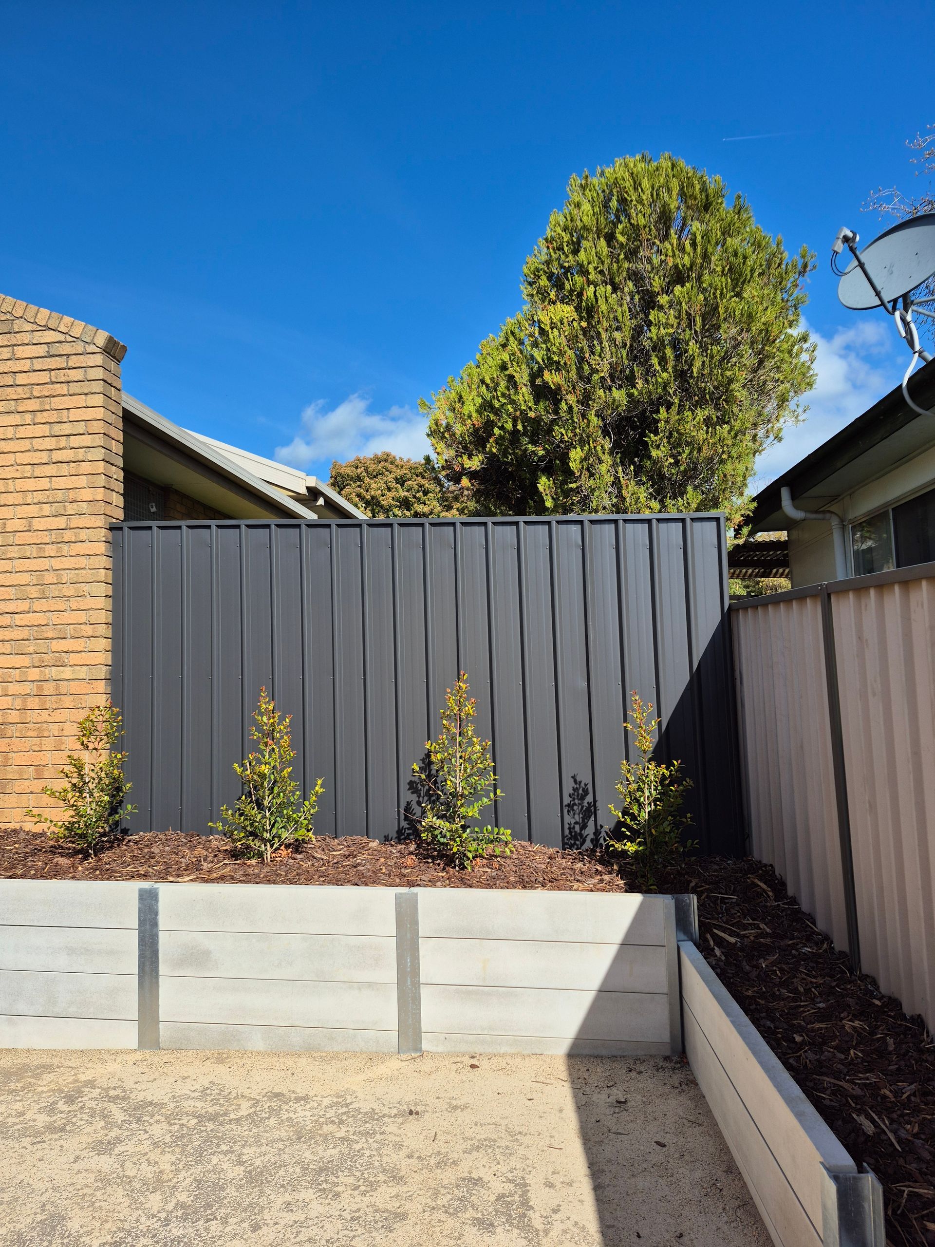 Gray vertical-slat fence with a retaining wall, small plants, and gravel ground under a blue sky — Precision Fencing & Landscaping in Thurgoona, NSW