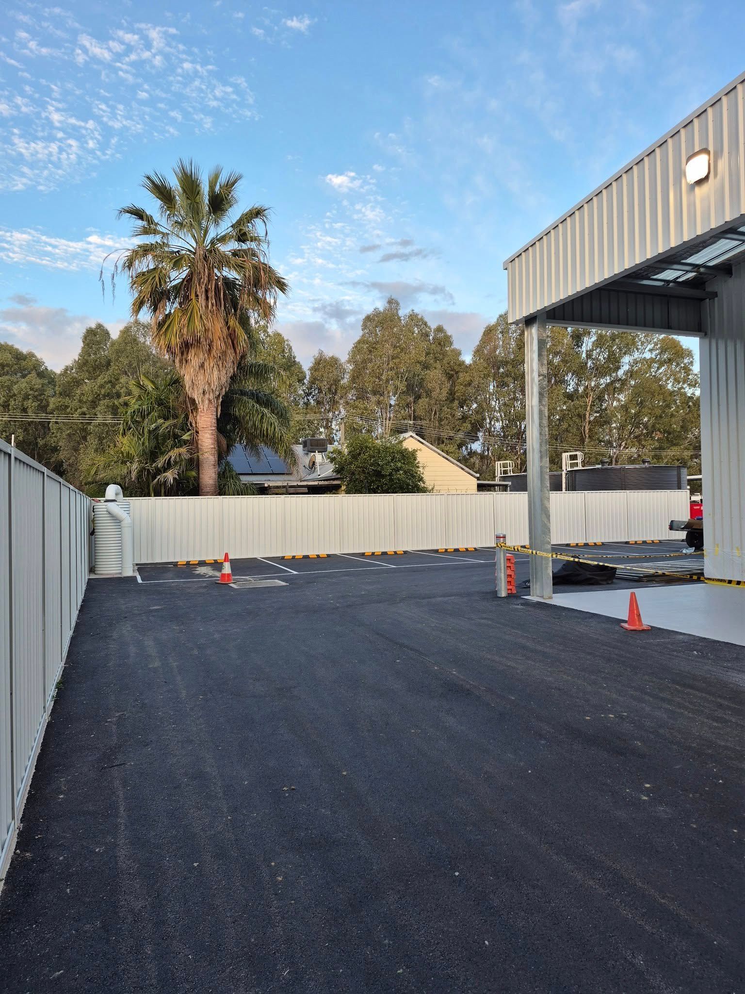 Gravel parking area next to a white fence and industrial building, with a tall palm tree in the background — Precision Fencing & Landscaping in Thurgoona, NSW